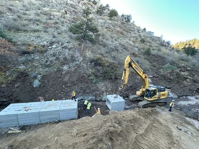 Construction workers and a backhoe working on placing large concrete blocks in a hilly area.