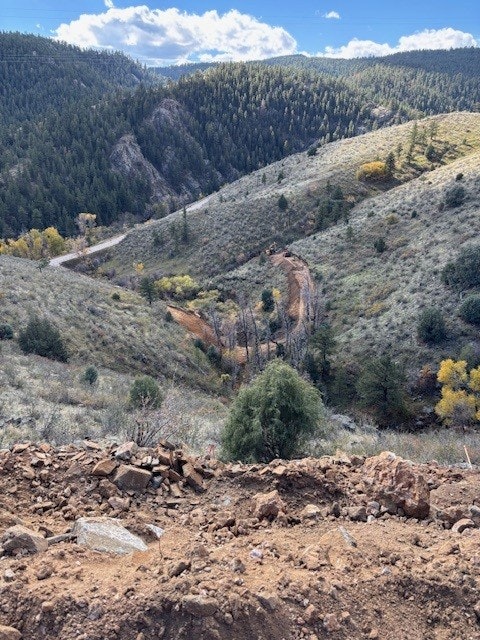 A dirt road winds through a hilly landscape covered with trees and shrubs under a blue sky with clouds.