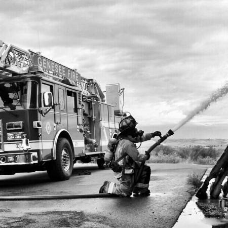 A firefighter spraying water from a hose next to a fire engine labeled "Genesee Fire." Black and white photo.