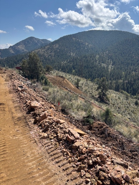 Mountain road construction with dirt tracks, rocks, and trees under a blue sky with clouds.