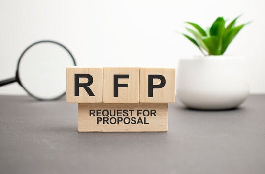 Wooden blocks with "RFP - Request For Proposal," a magnifying glass, and a potted plant on a desk.