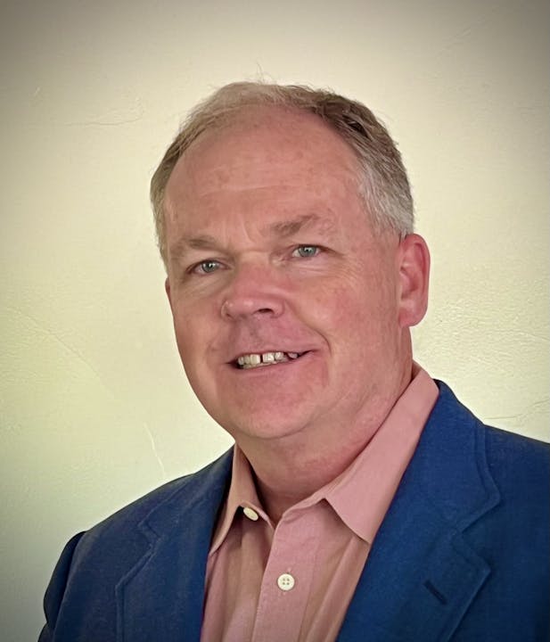 A person smiling, wearing a blue jacket and pink shirt, against a plain background.