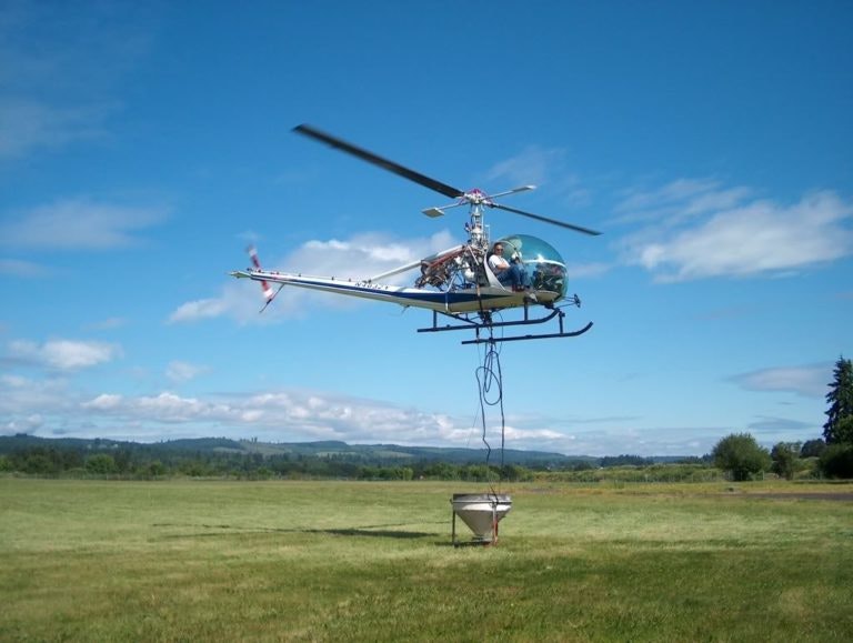 A helicopter hovers above a grassy field, holding a large container below it with cables.