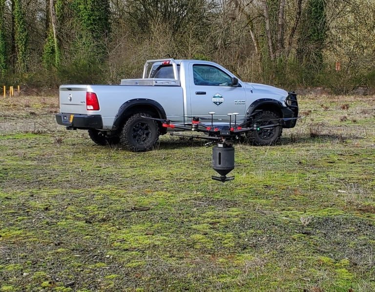 A silver pickup truck is parked in a field with a large drone nearby on the ground.