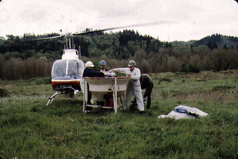 A group of people working near a helicopter in a grassy field, with trees in the background.