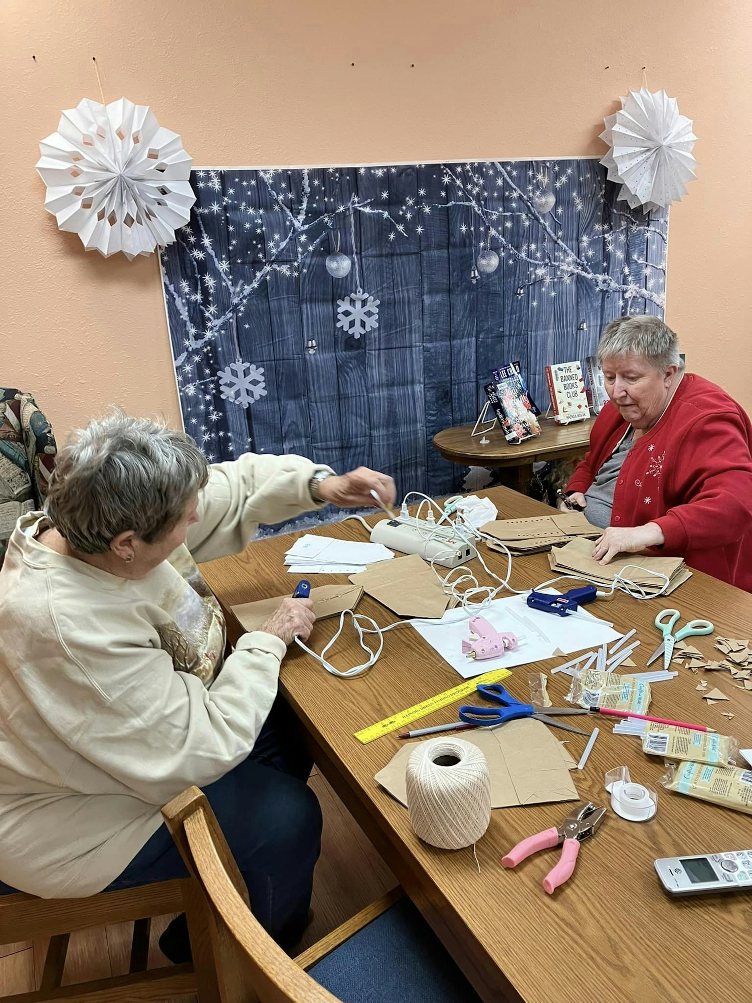 Two people are crafting at a table with paper and tools. Snowflake decorations and winter backdrop are visible.