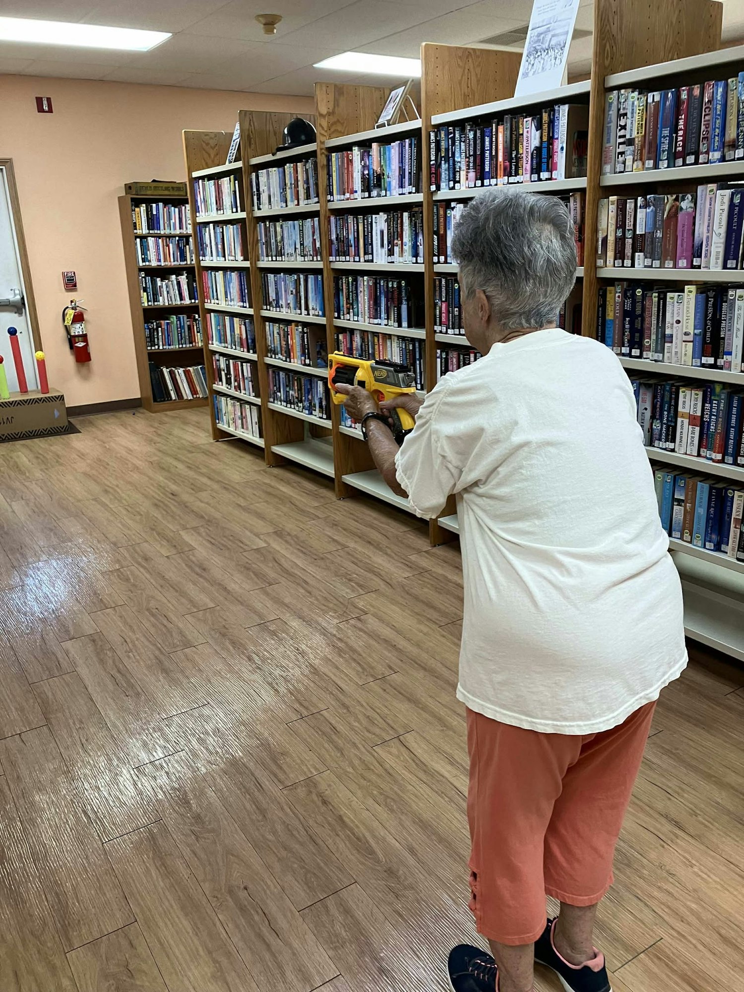 An older woman in a library aiming a toy gun, standing on a wooden floor.