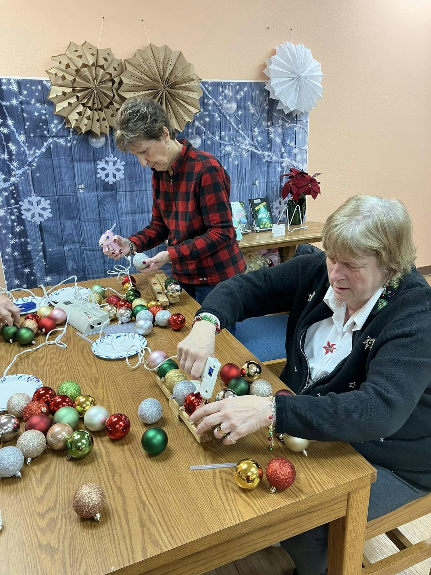 Two people crafting with Christmas ornaments on a table decorated with festive paper rosettes and lights.