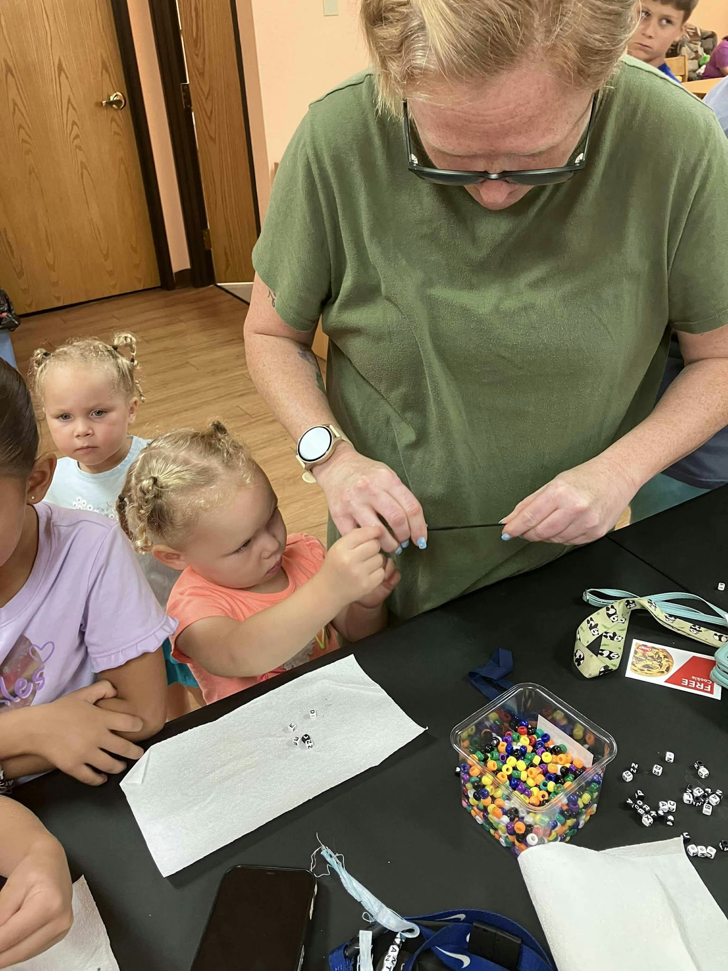 A group engaged in a beading activity; an adult assists a child while others watch.