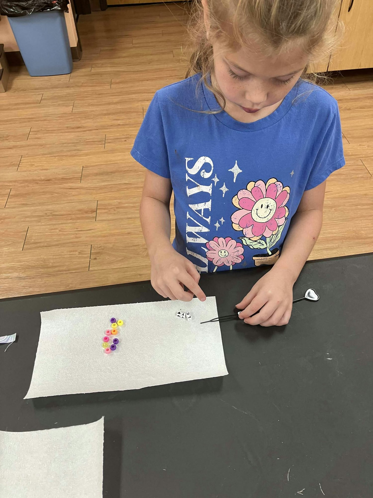 A child in a blue floral shirt is arranging beads on a paper sheet using a stick with a heart-shaped end.