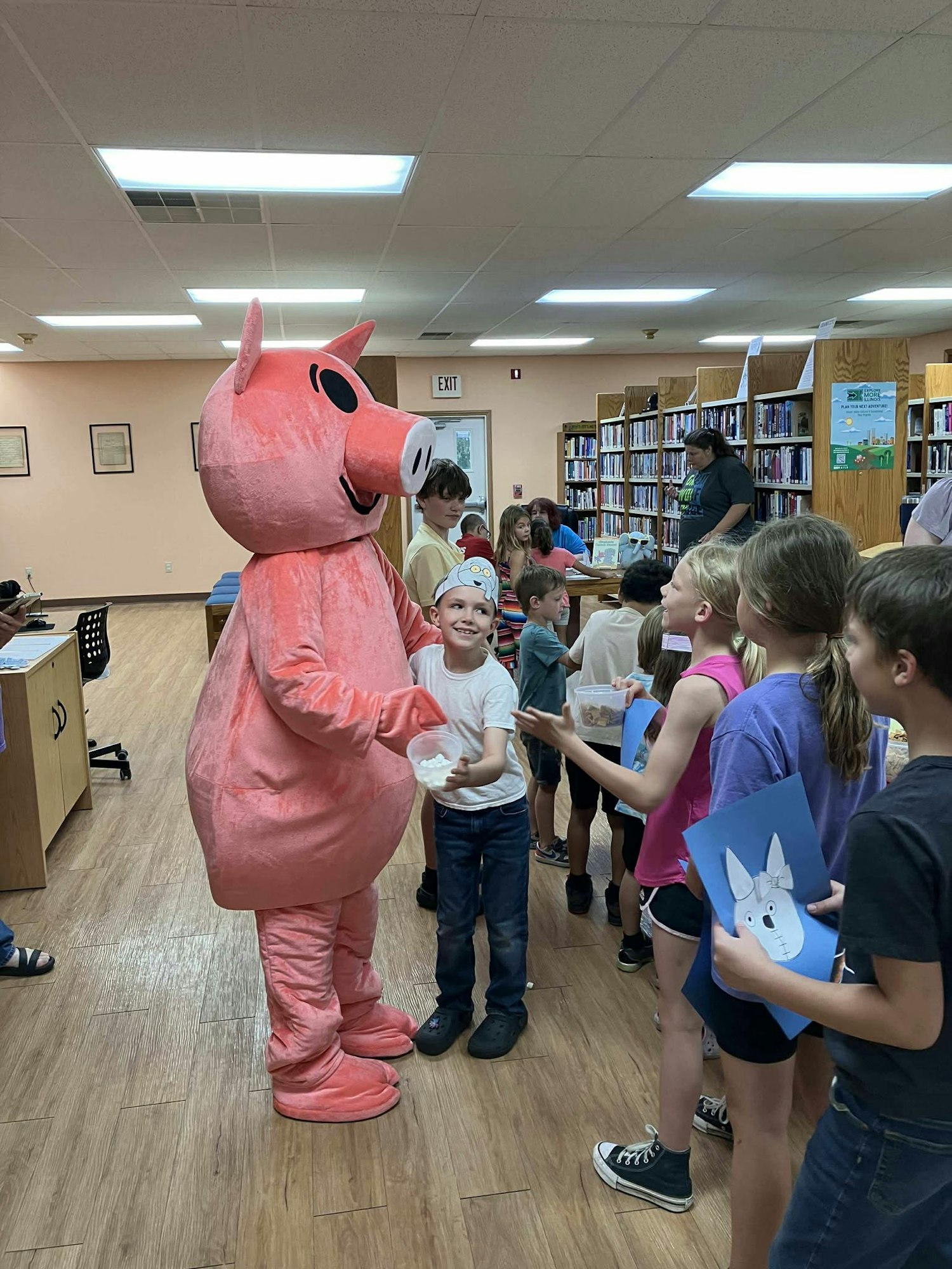 A person in a pig costume interacts with a group of children in a library.
