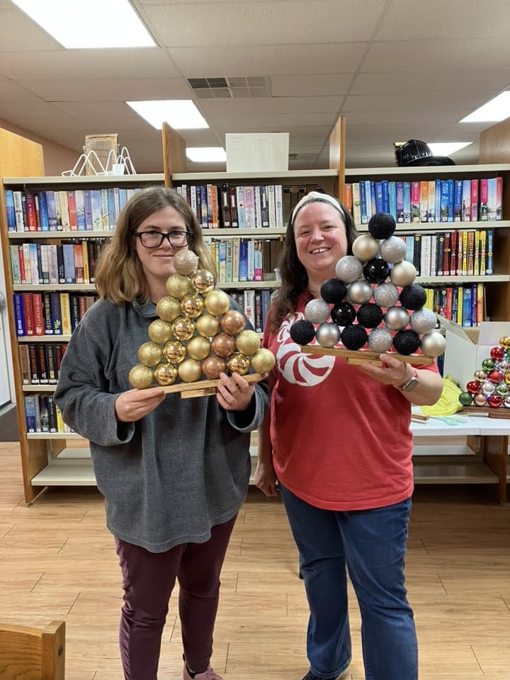 Two people holding triangular arrangements of decorative orbs in front of a bookshelf.