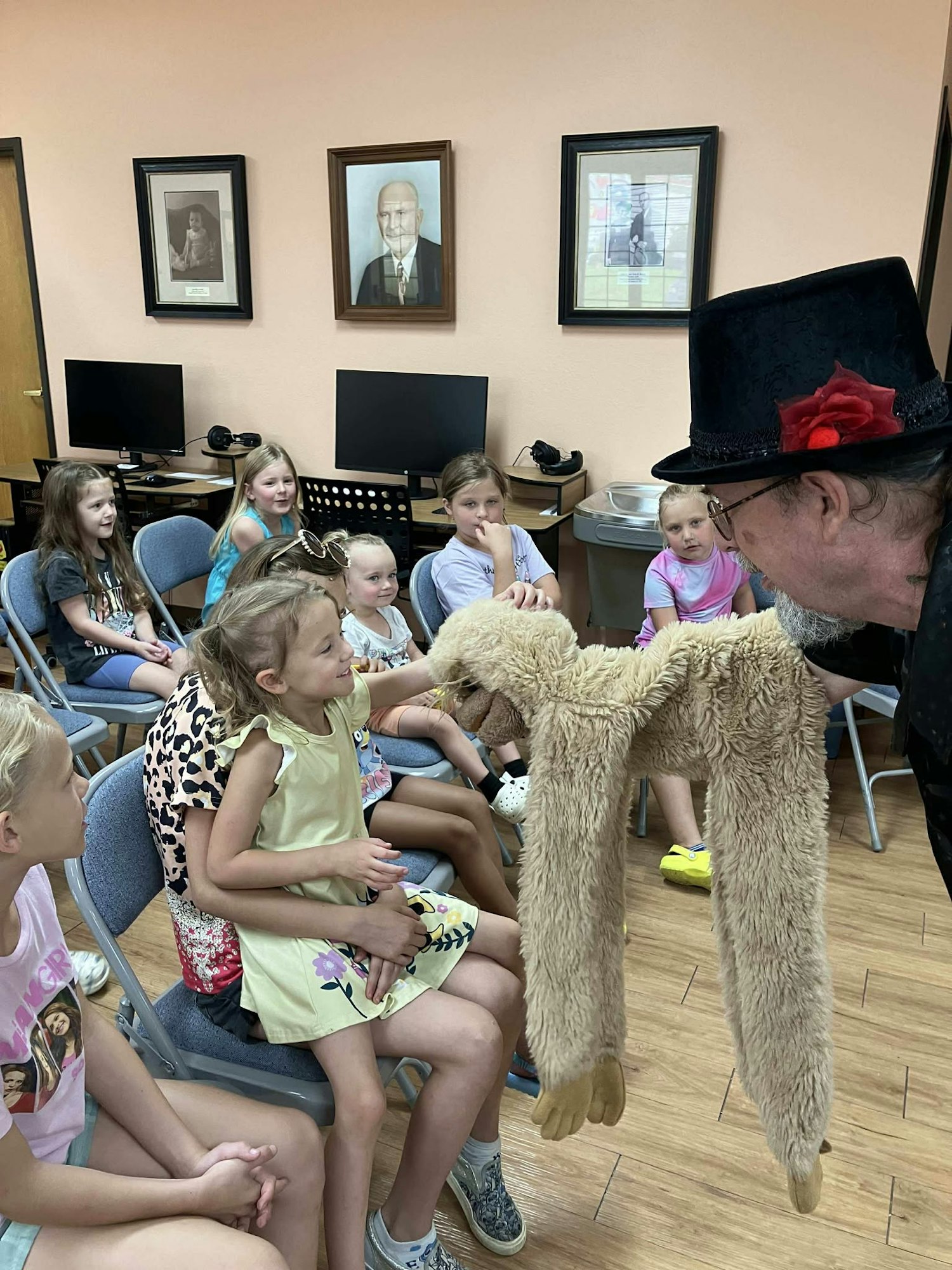 A man in a hat shows a puppet to a group of children seated in a room with framed photos on the wall.