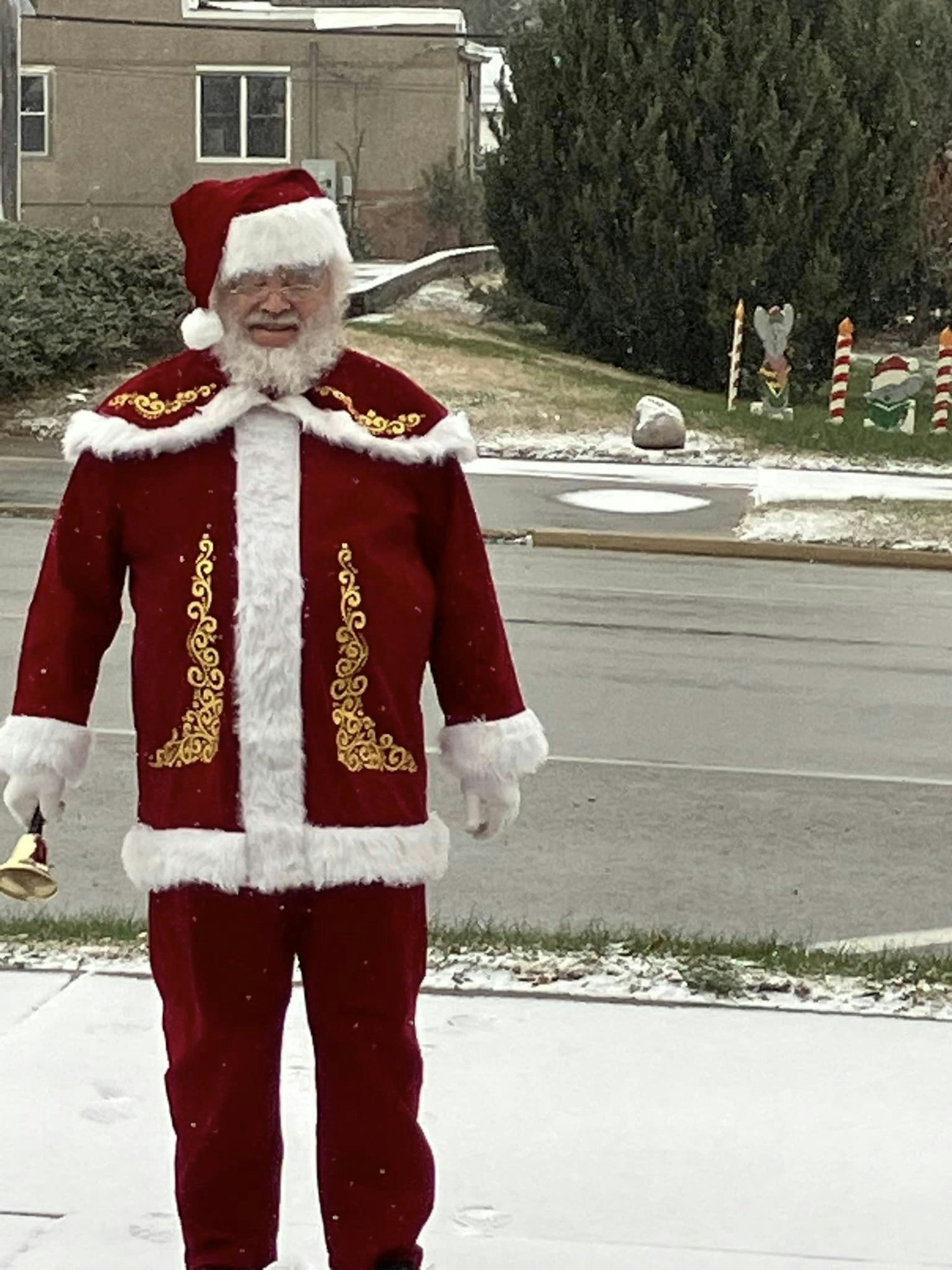 A person dressed as Santa Claus standing on a snowy sidewalk, holding a bell.