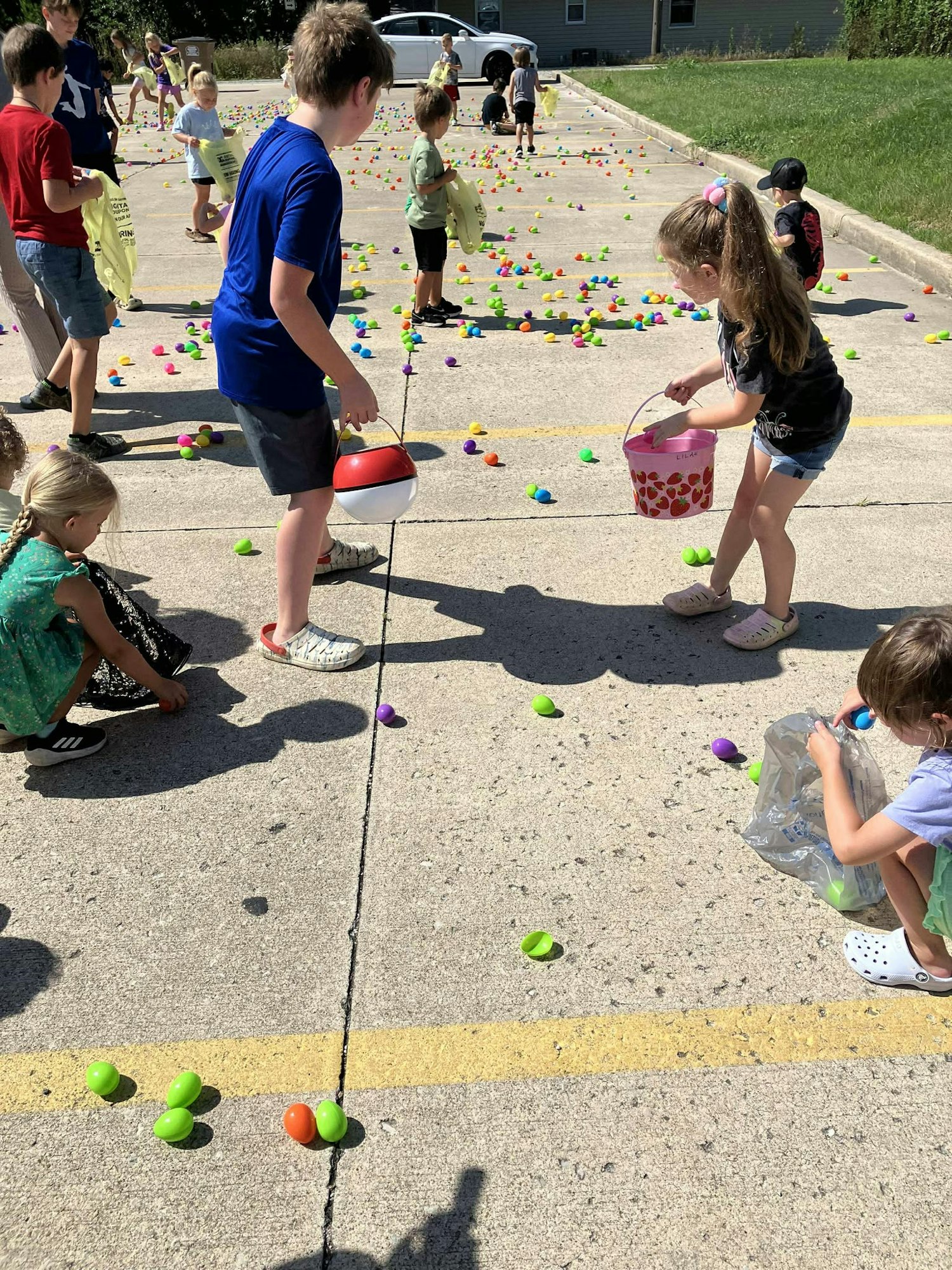 Children collecting colorful eggs scattered on a pavement, using baskets and bags.