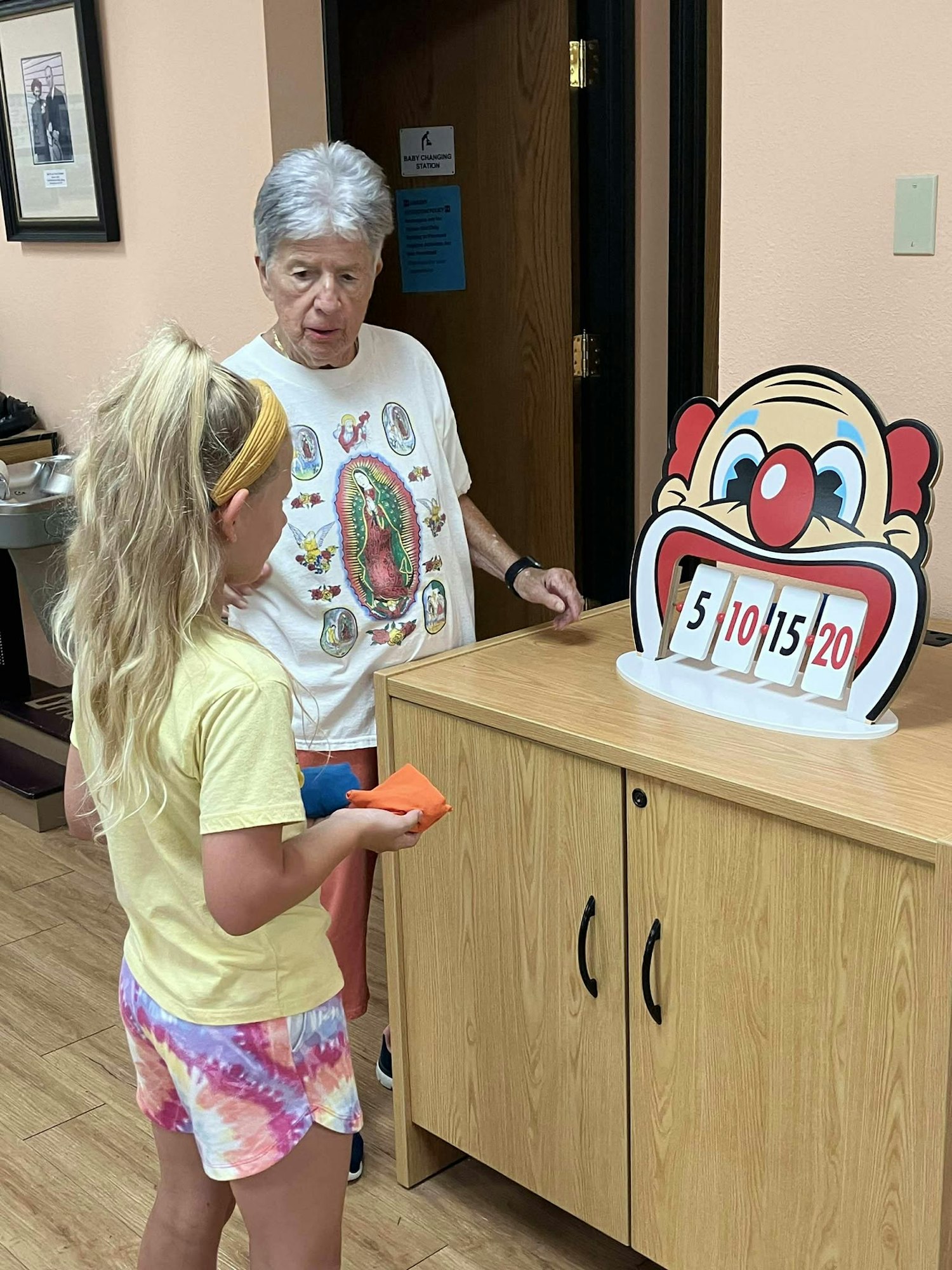 A child and an older person are playing a game with a clown face target showing scores 5, 10, 15, 20, on a cabinet.