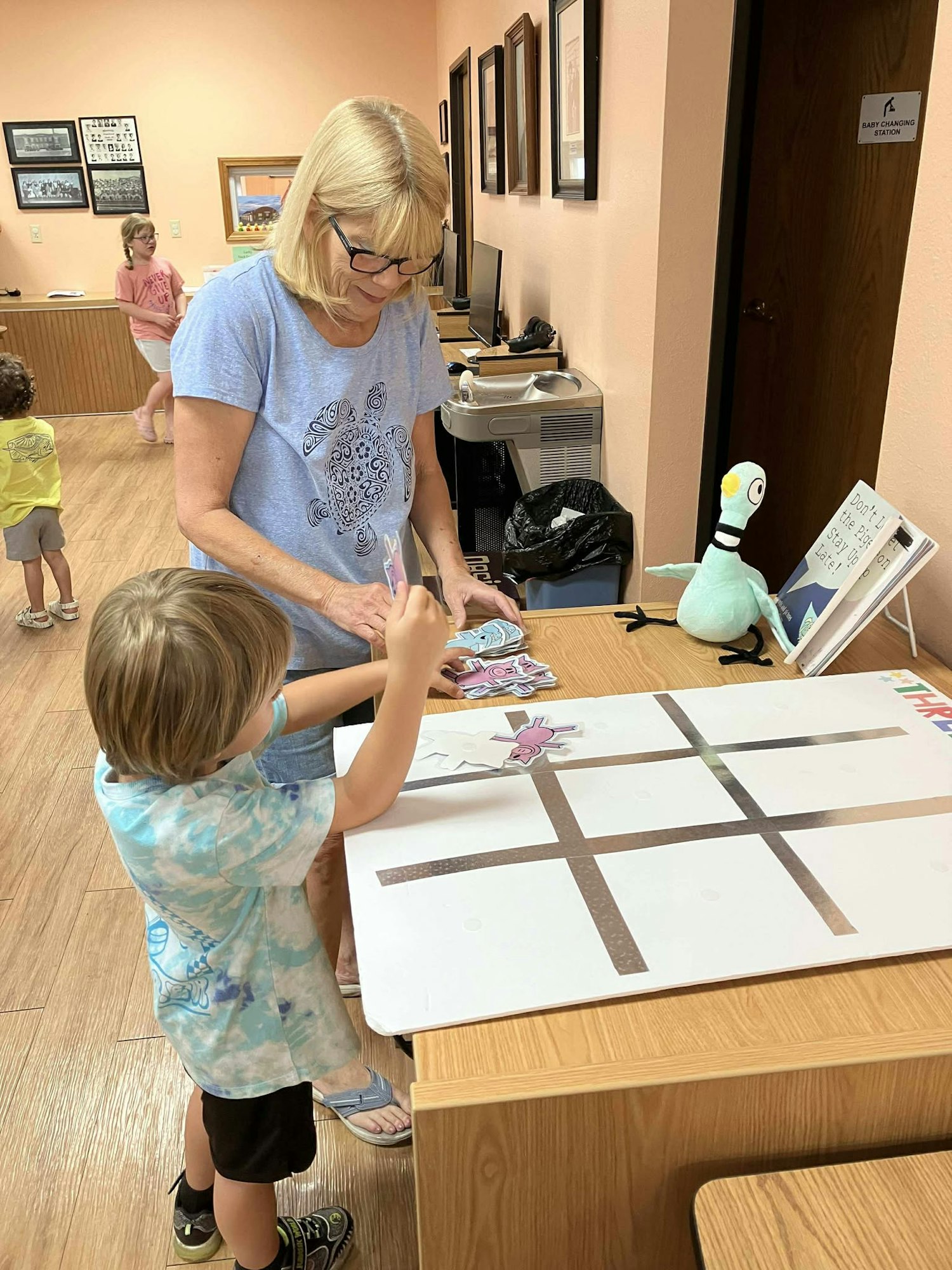 A woman and child work on a tic-tac-toe game at a table indoors. Children are in the background. Nearby is a plush toy.