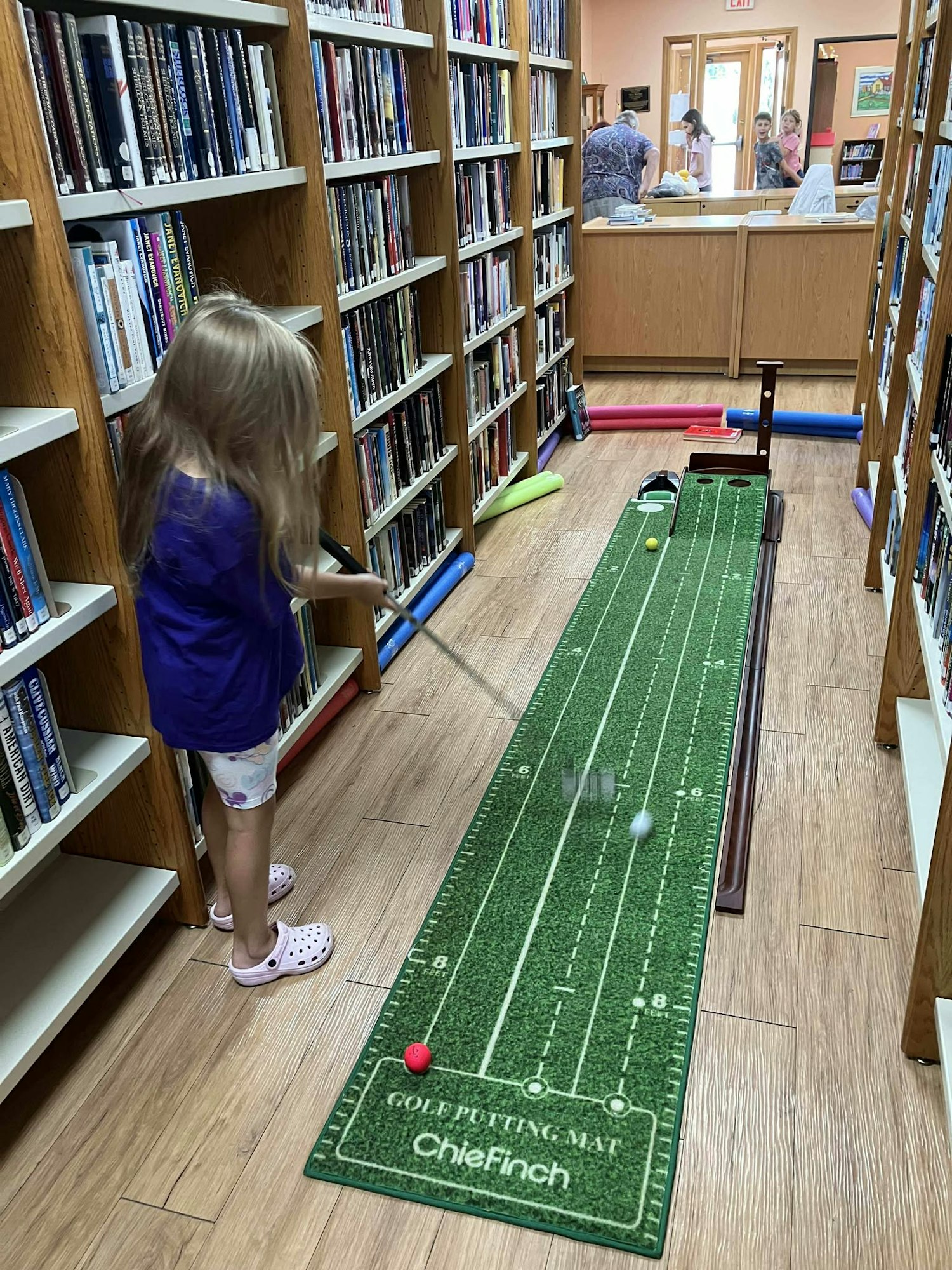 A girl playing mini-golf on a putting mat in a library aisle.