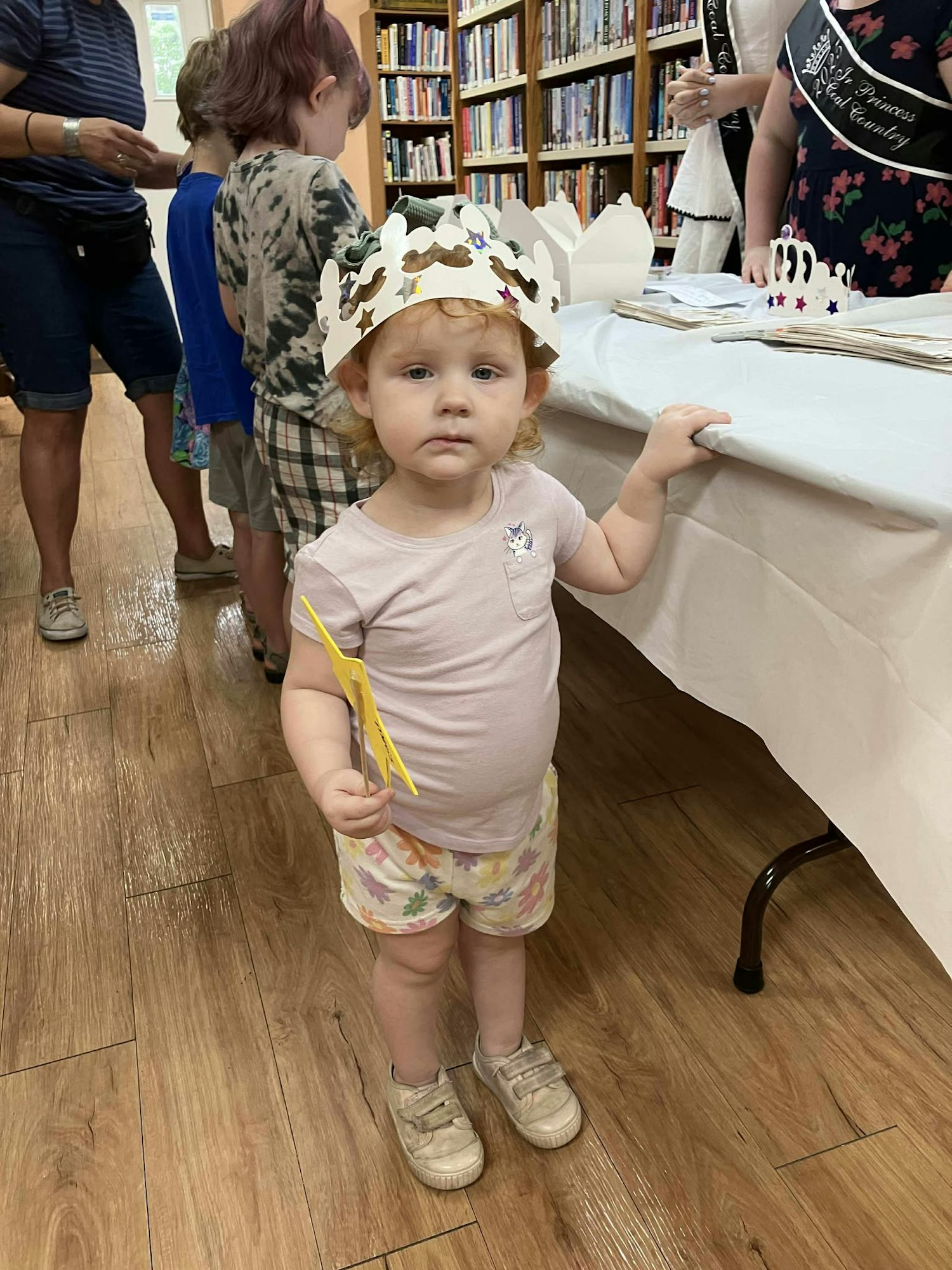 A child wearing a paper crown stands in a library beside a decorated table, holding a crafting tool.