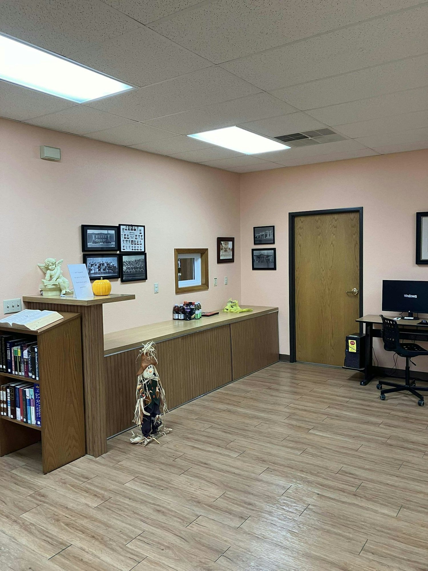 A tidy office interior with a reception desk, computer, shelving with books, decorative scarecrow, and framed photographs on the wall.