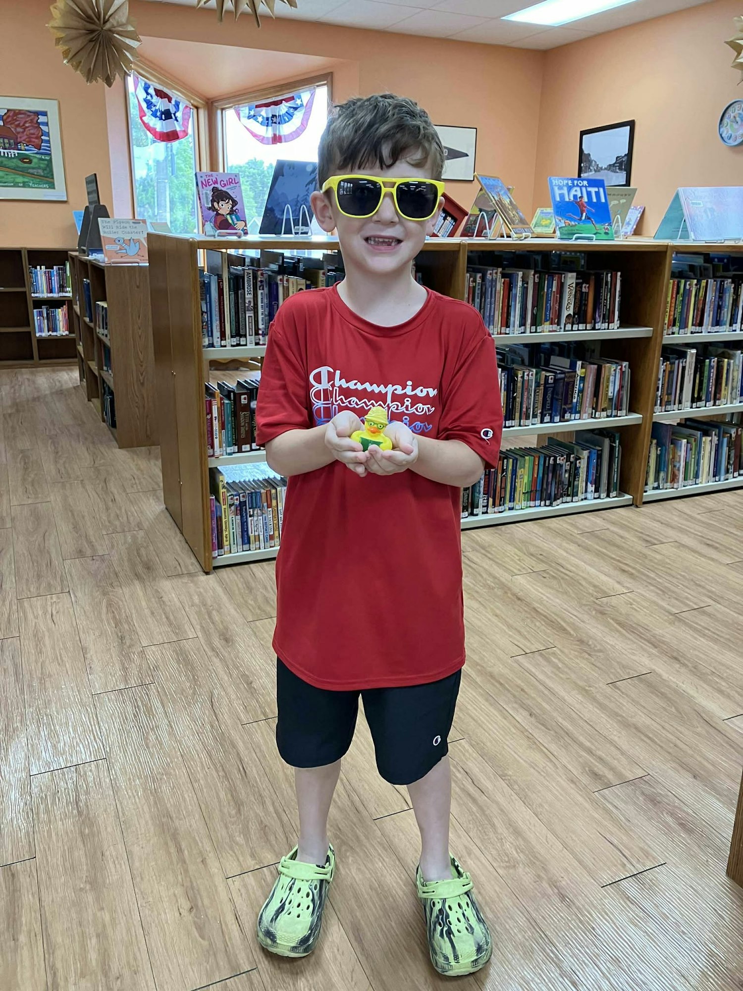 A child in sunglasses holding a toy in a library, wearing a red shirt and green shoes.