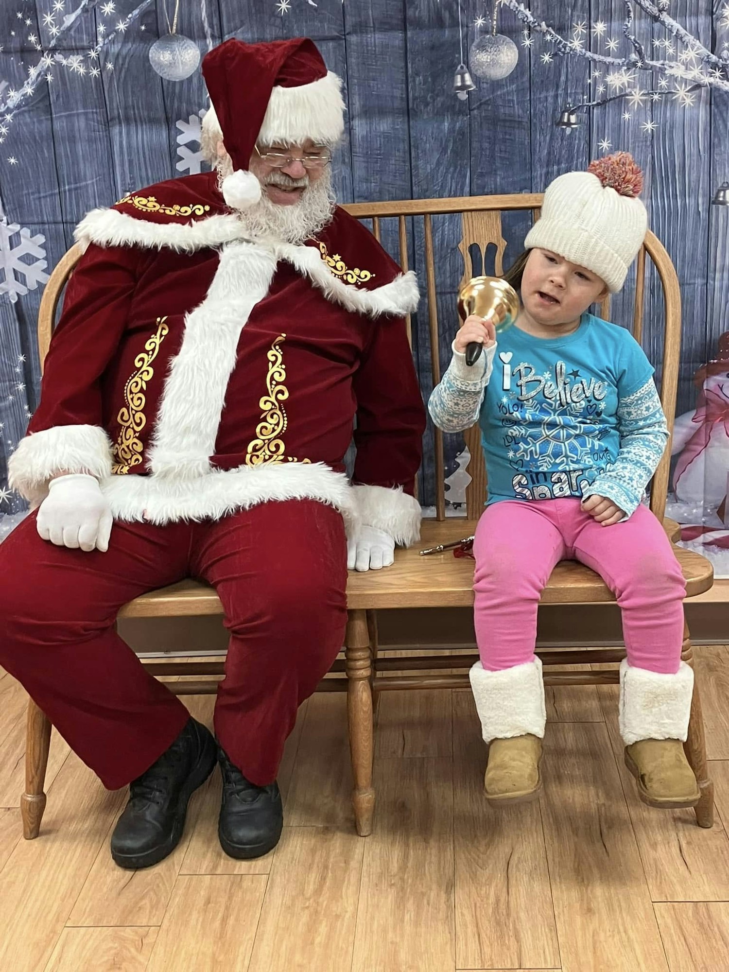 Santa sitting on a bench with a child in a festive setting, holding a bell.