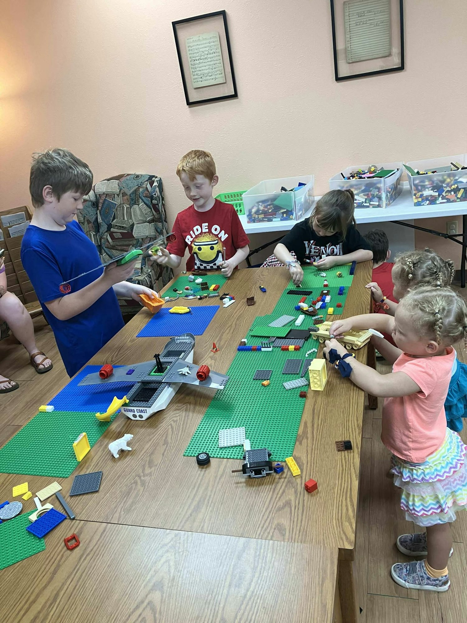 Children building with LEGO pieces on a table, surrounded by bins of bricks.