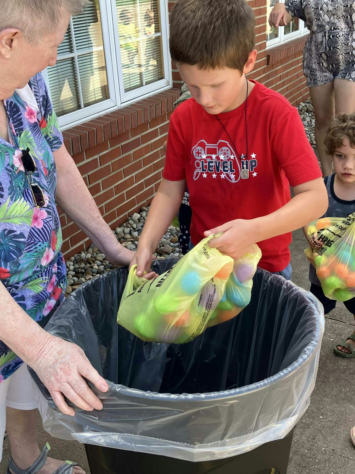 A child in a red shirt places a bag of colorful eggs into a trash bin, assisted by an adult.
