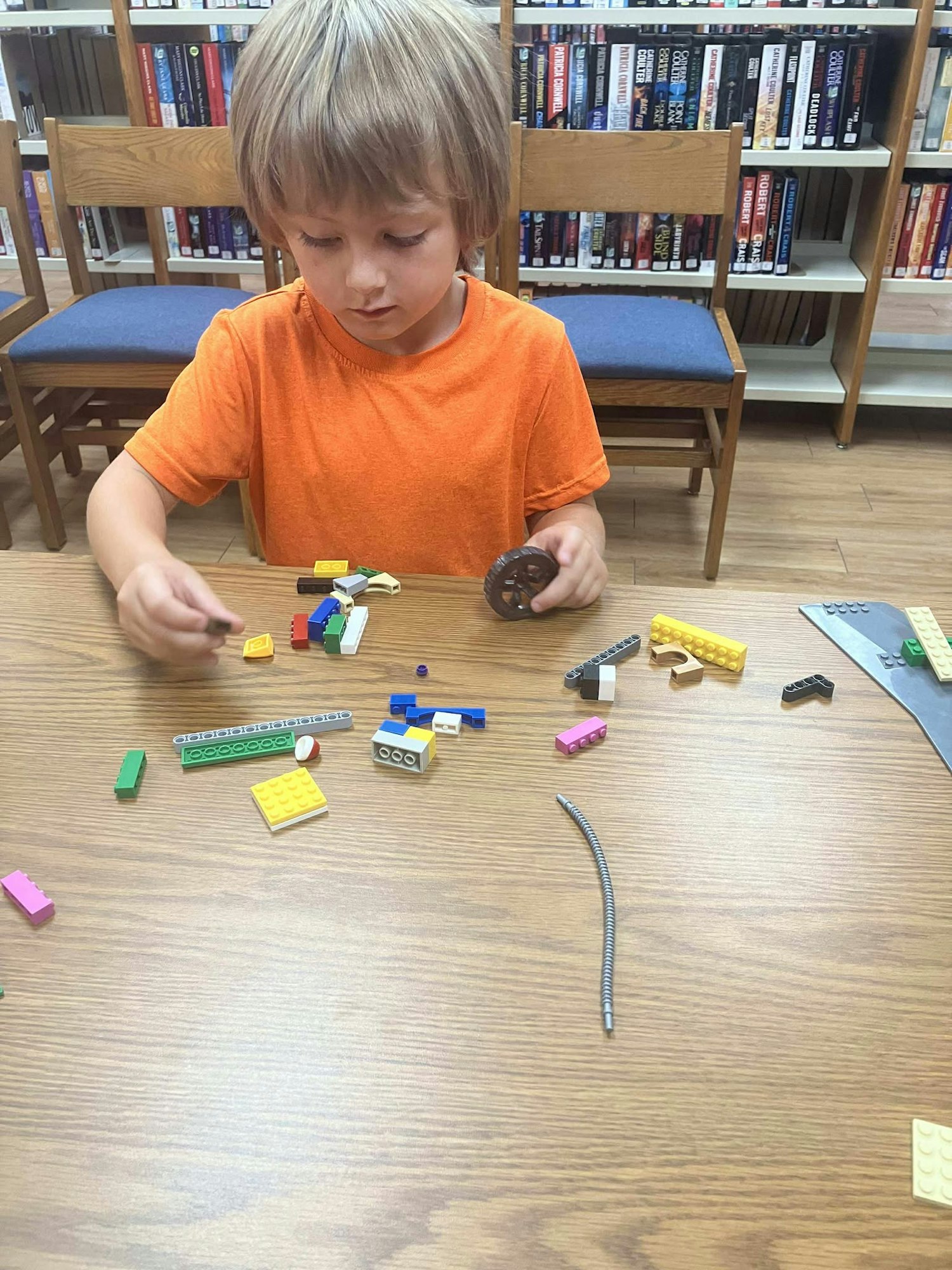 A child in an orange shirt is building with LEGO bricks on a wooden table in front of bookshelves.