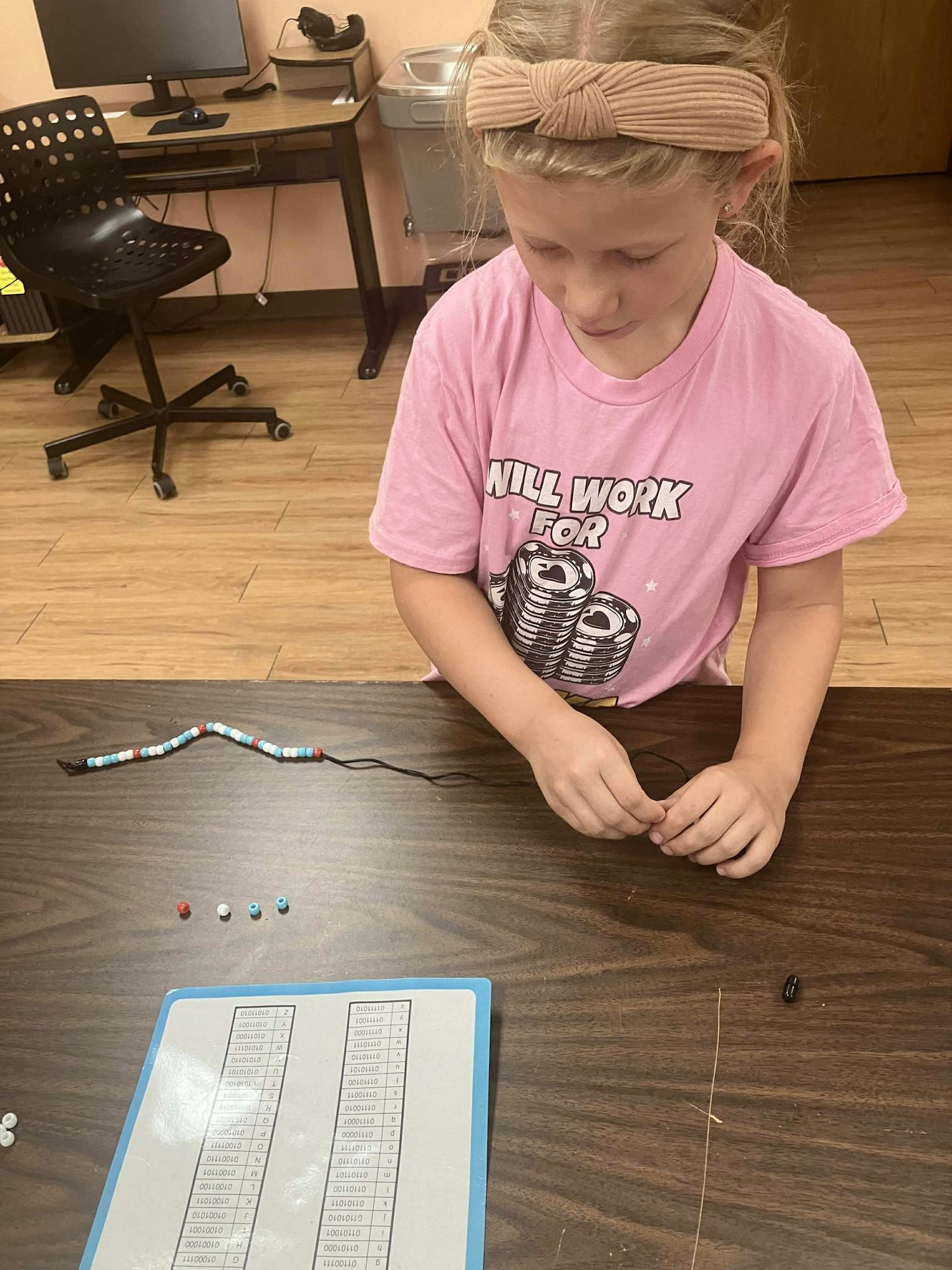 Child making a bracelet with colorful beads at a table, next to a binary alphabet sheet and a computer in the background.