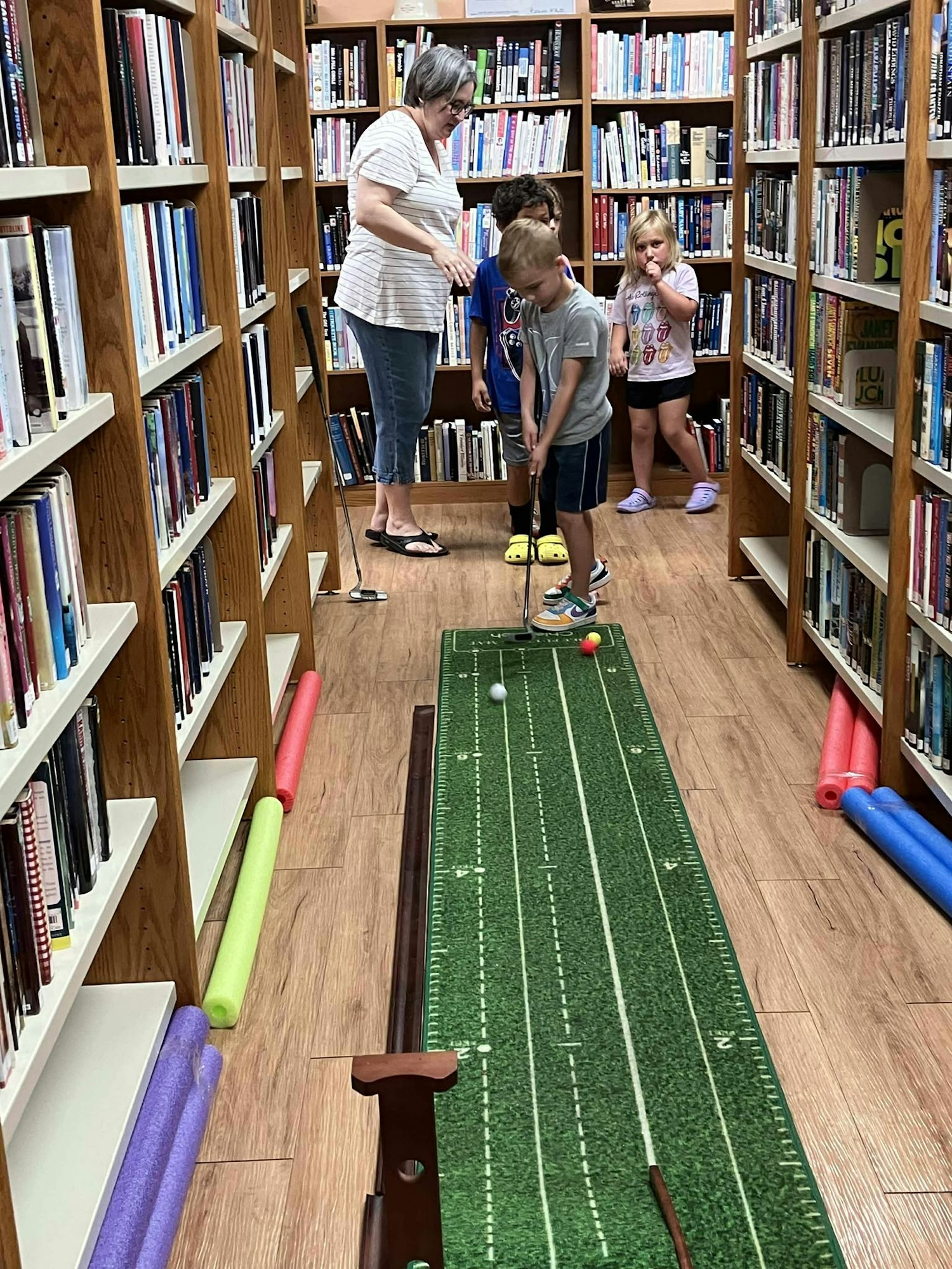 Children playing mini-golf in a library aisle, guided by an adult, surrounded by bookshelves.