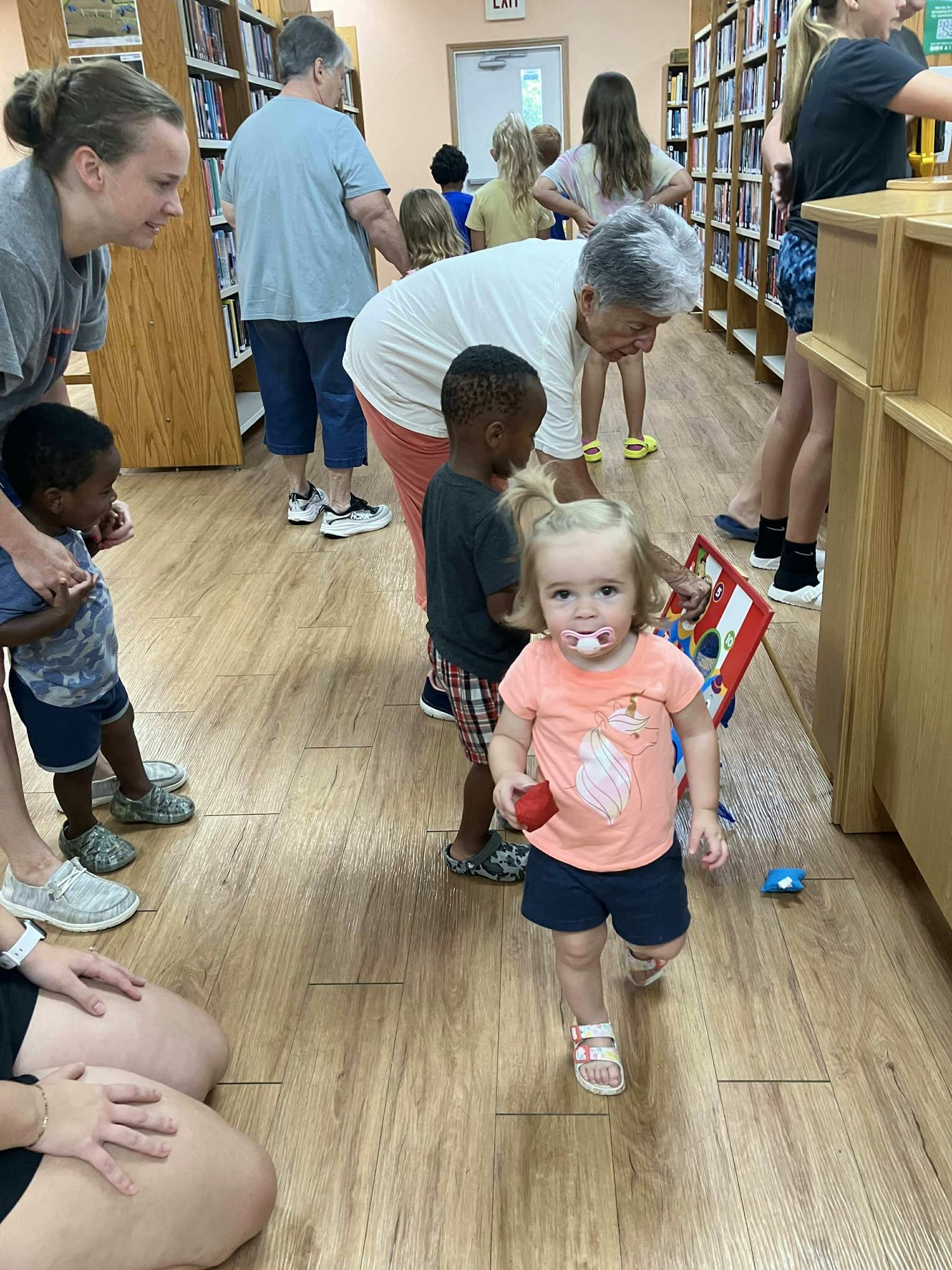 Children and adults in a library, some reading and interacting, one toddler walking towards the camera.