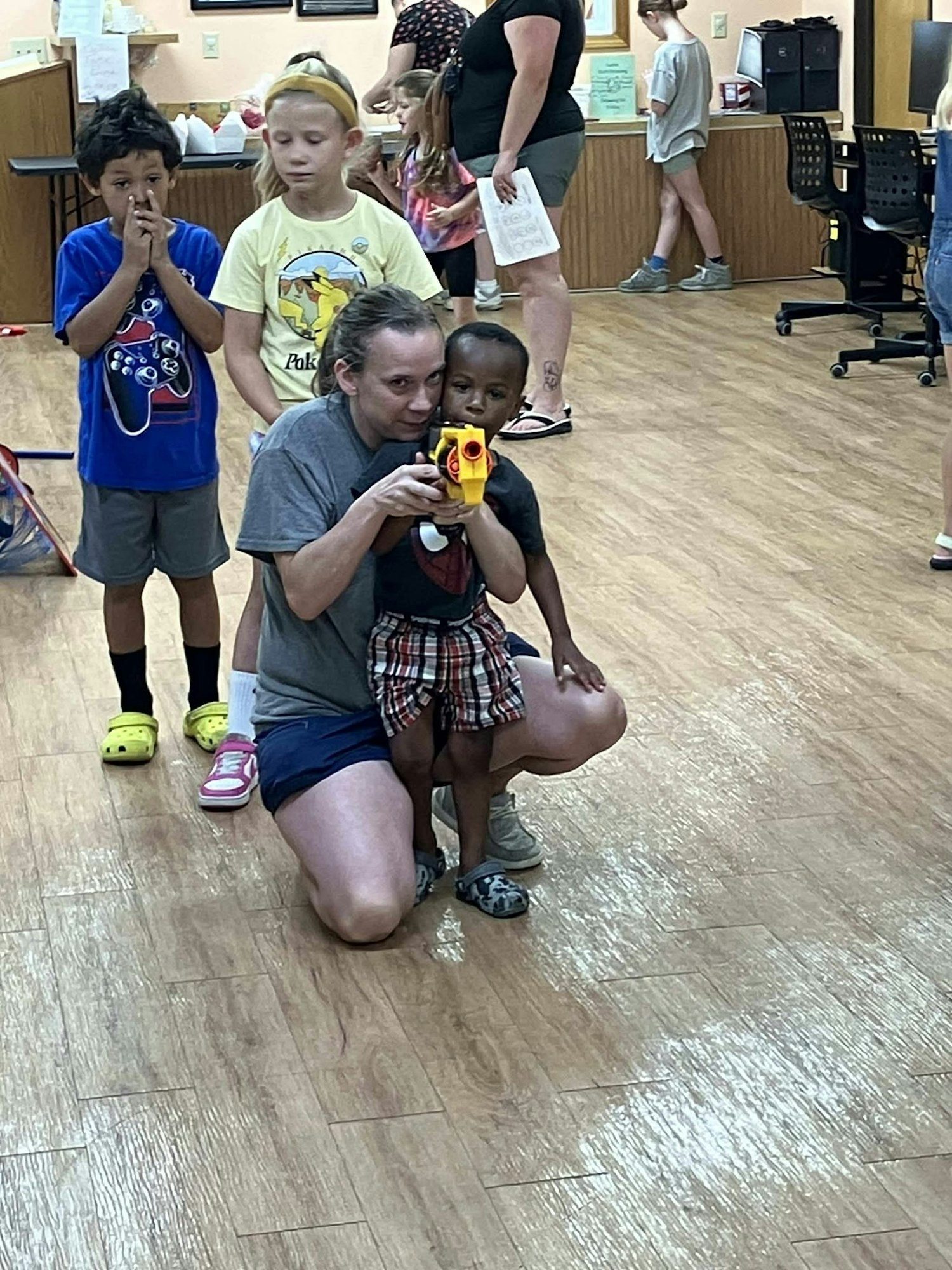 A woman kneels and assists a child aiming a toy gun, while other children and adults are in the background indoors.