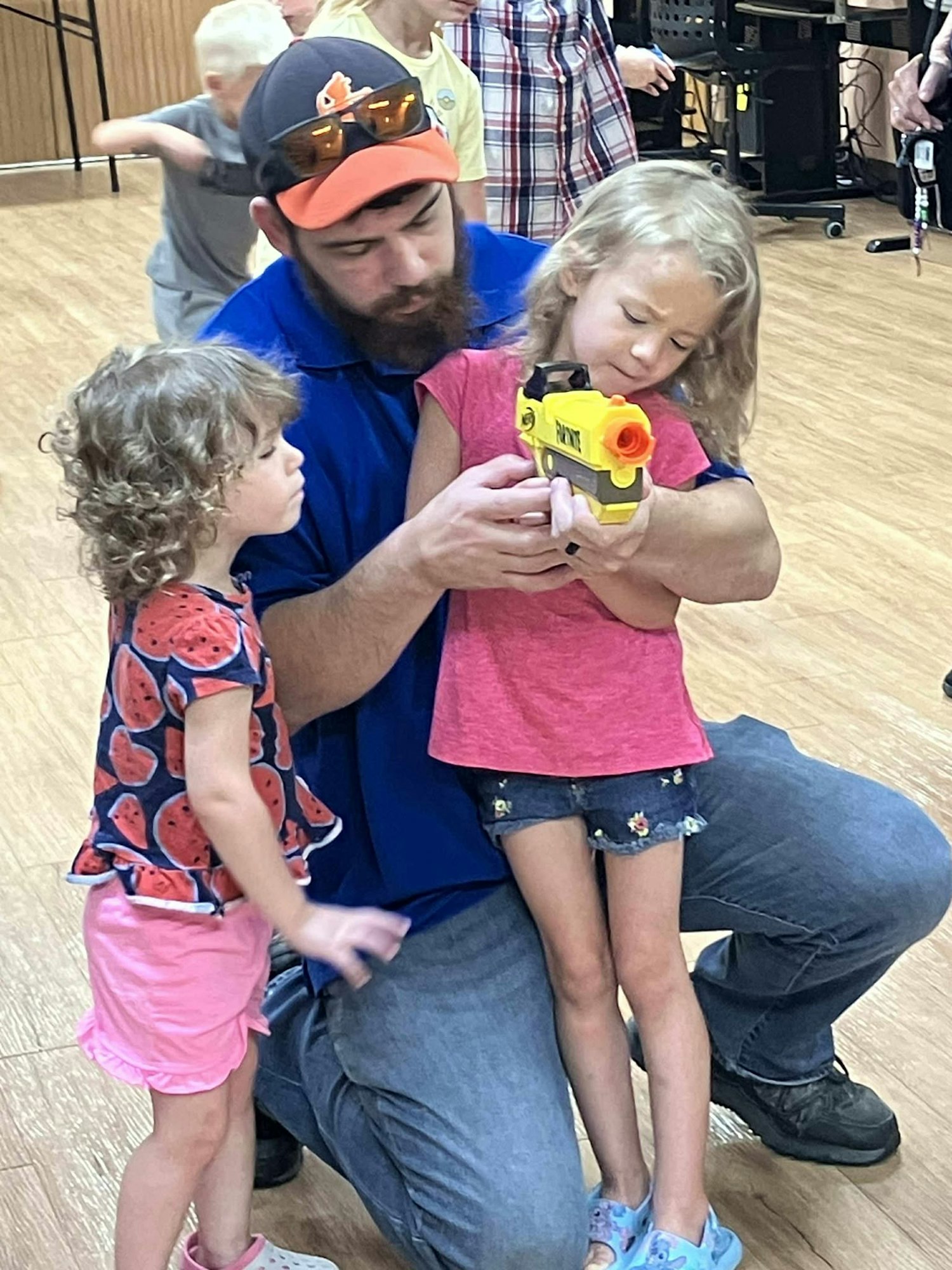 A man kneels, helping a child hold a toy gun, while another child watches.