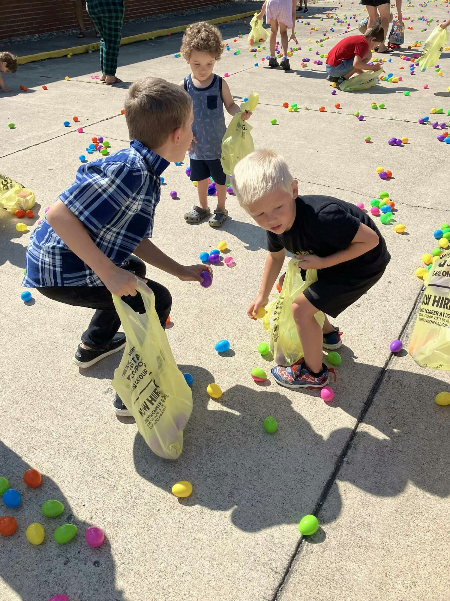 Children collecting colorful plastic eggs outdoors with yellow bags.