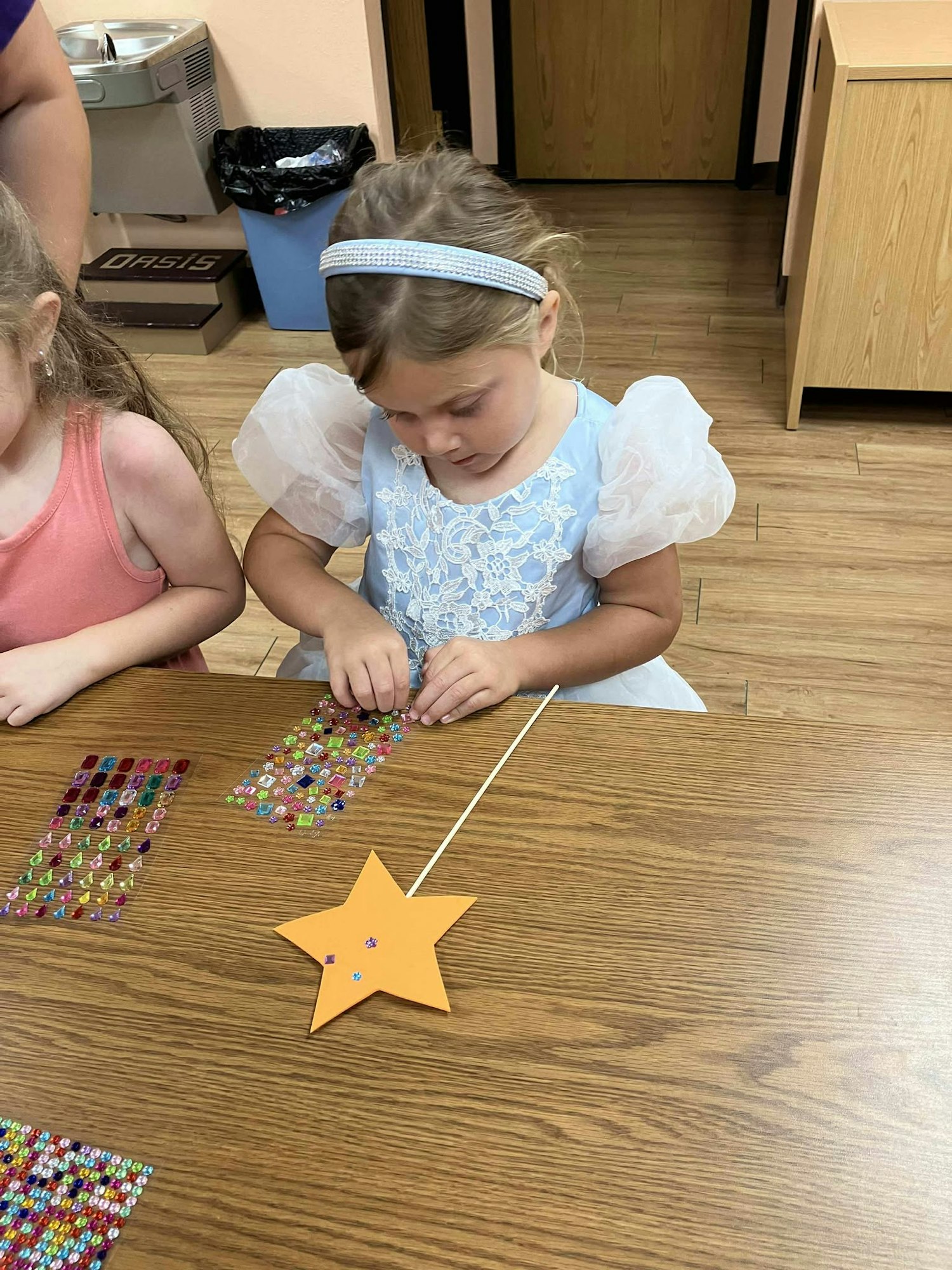 A child in a light blue dress decorates a star-shaped wand with colorful stickers at a table.