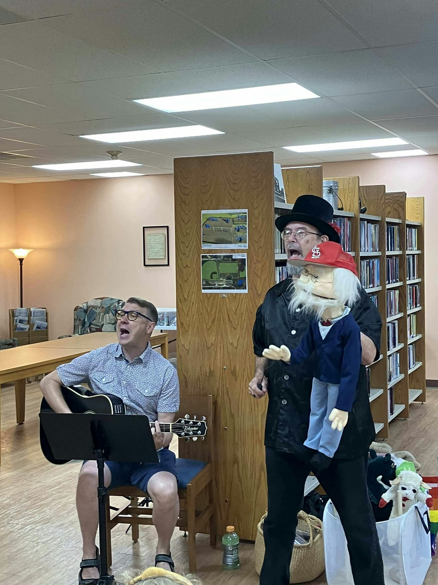 A person plays guitar while another, with a puppet, performs in a library setting.