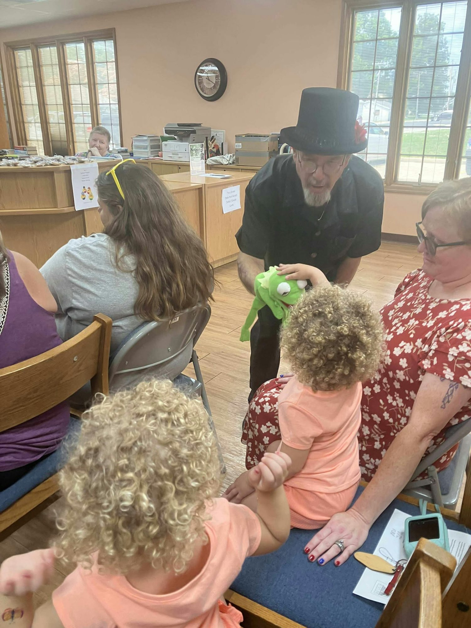A man in a hat entertains two children with a green puppet in a room with other people seated.