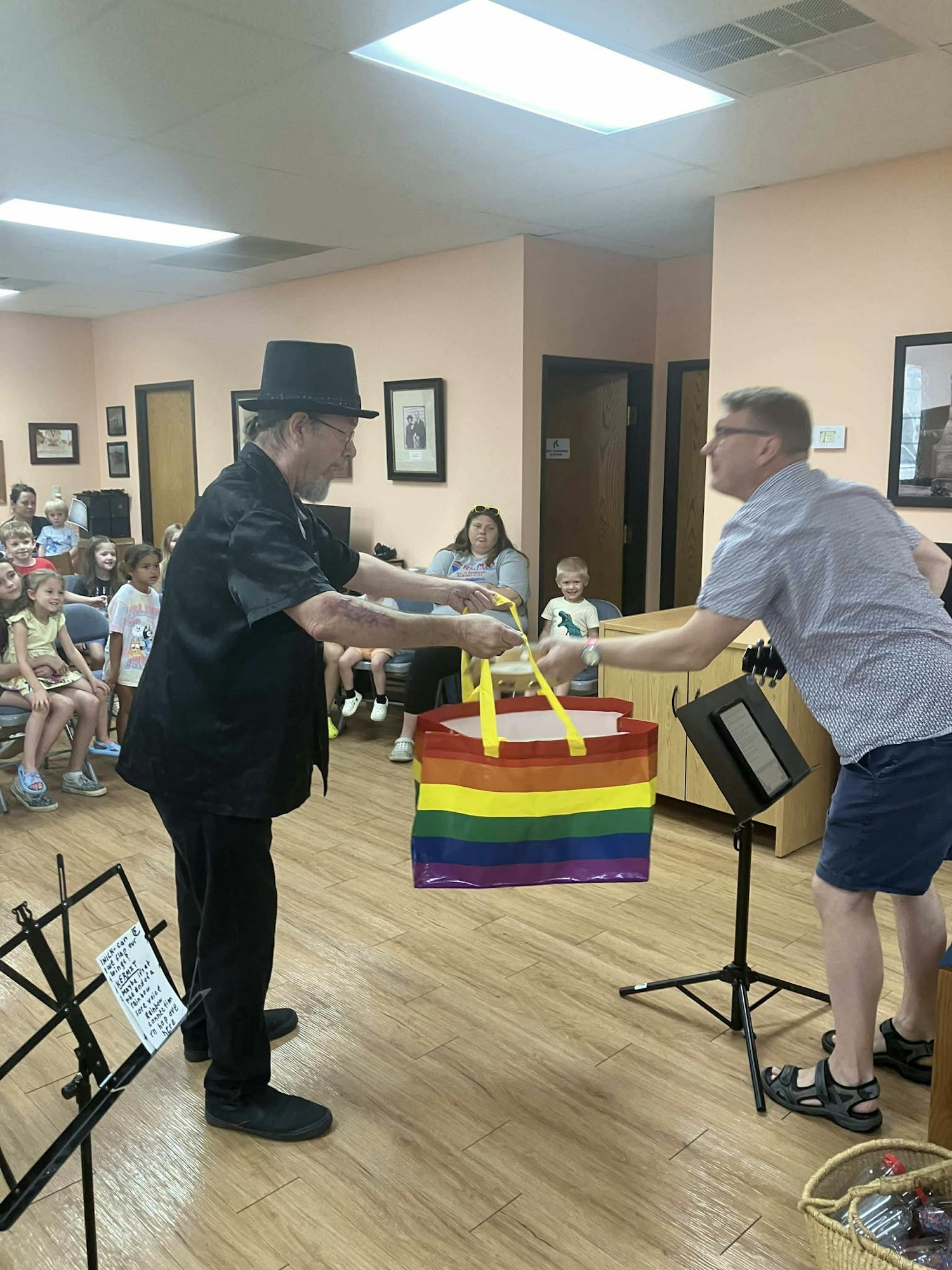 A man in a hat hands a rainbow-striped bag to another man in a room with seated children watching.