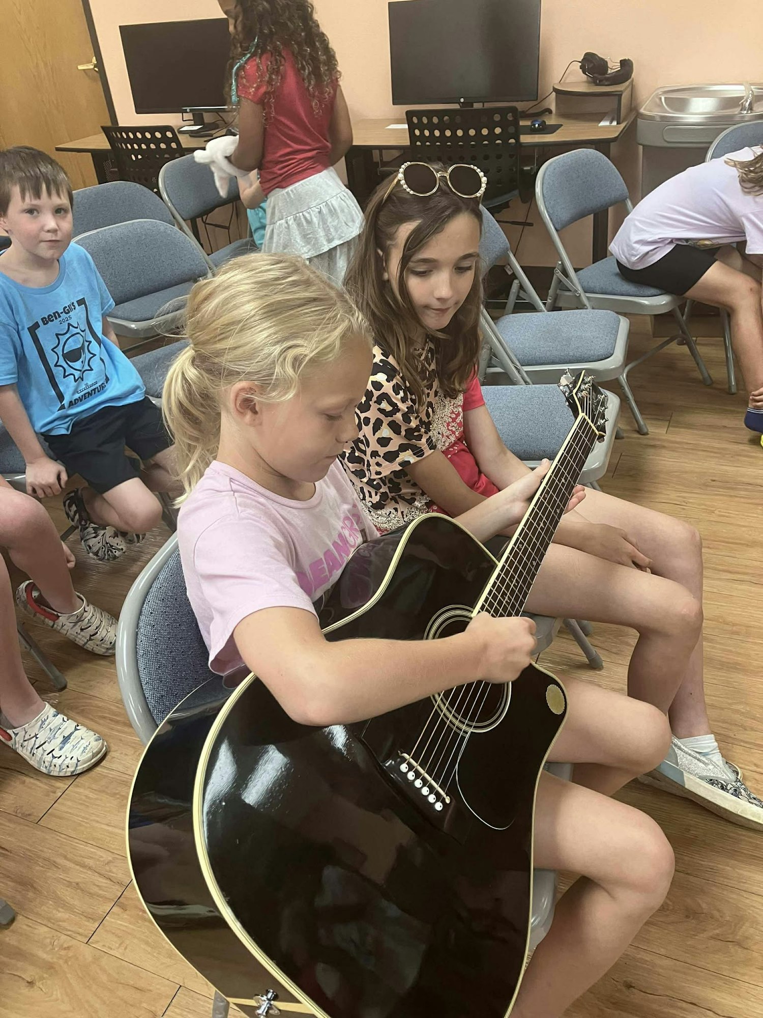 Kids sitting in a room, one playing a black acoustic guitar, others watching.