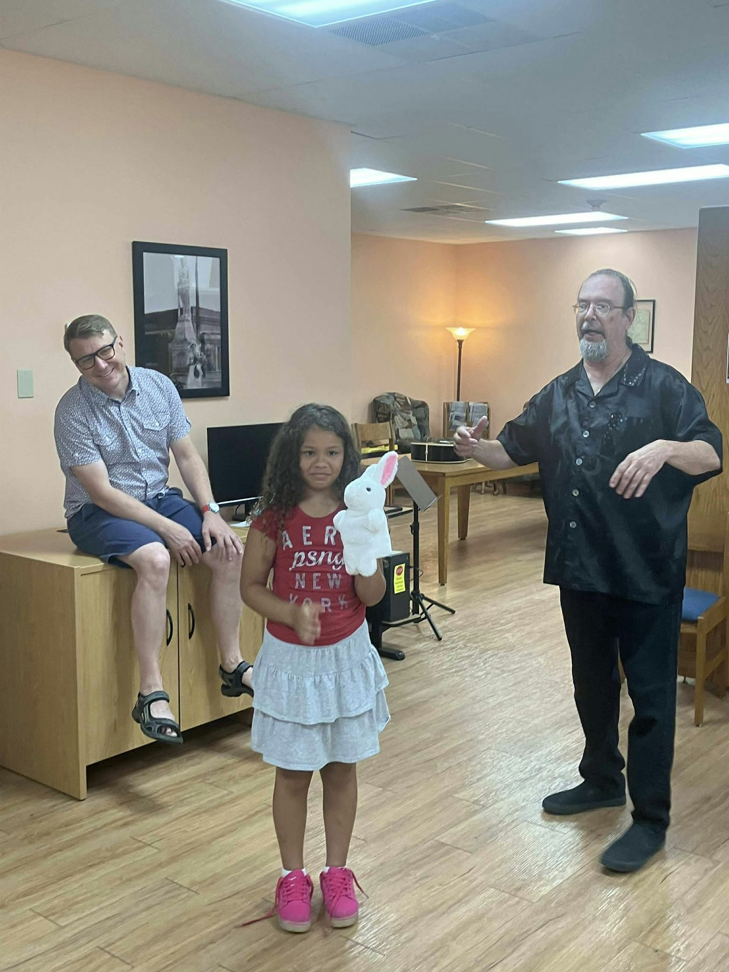 A girl holds a plush toy in a room with two men, one seated and the other gesturing.