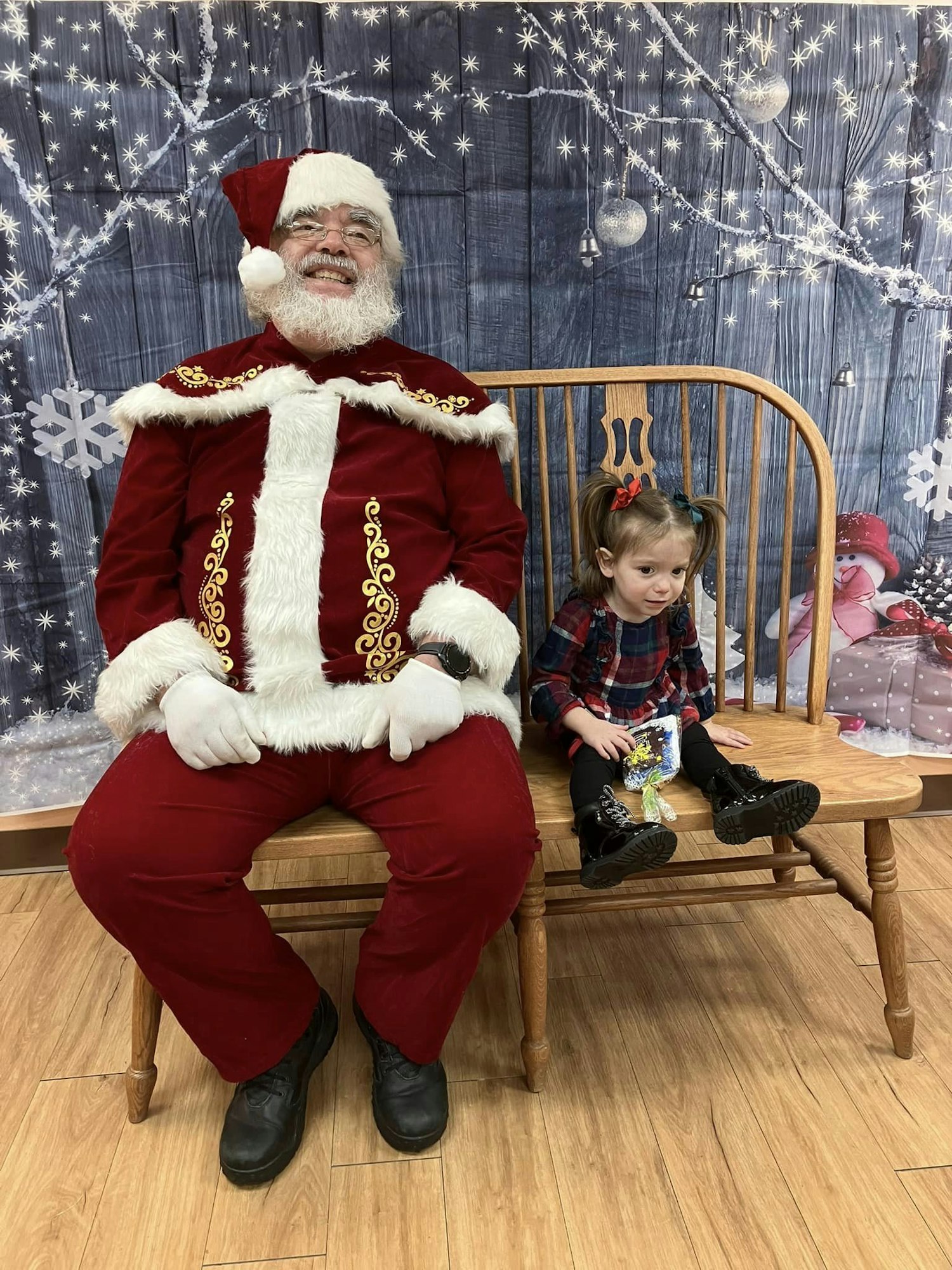 Santa Claus sits on a bench next to a little girl holding a gift, with festive decorations in the background.