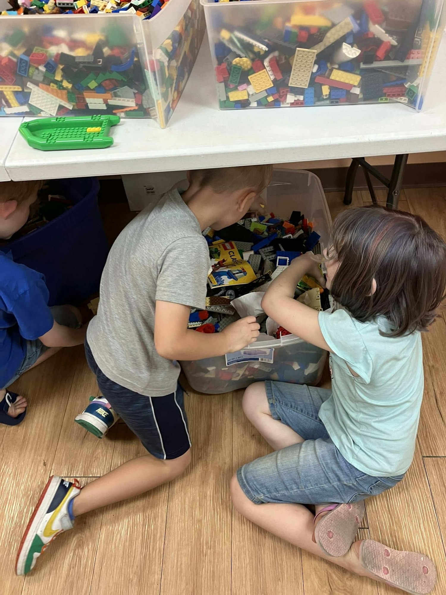 Children playing with LEGO blocks in a room, surrounded by various bins of colorful pieces.