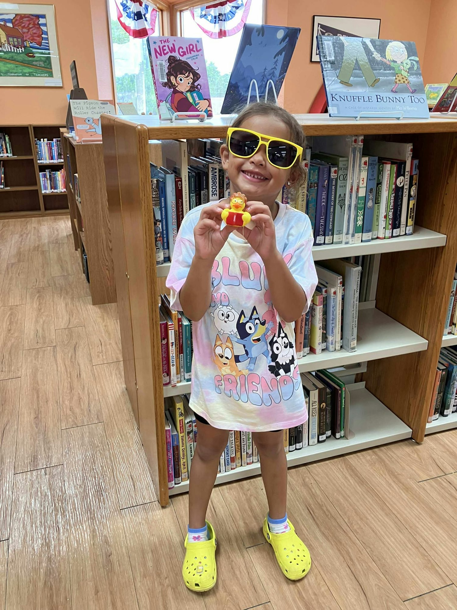 Child in a library wearing sunglasses and yellow Crocs, holding a toy; shelves filled with books and displays in the background.