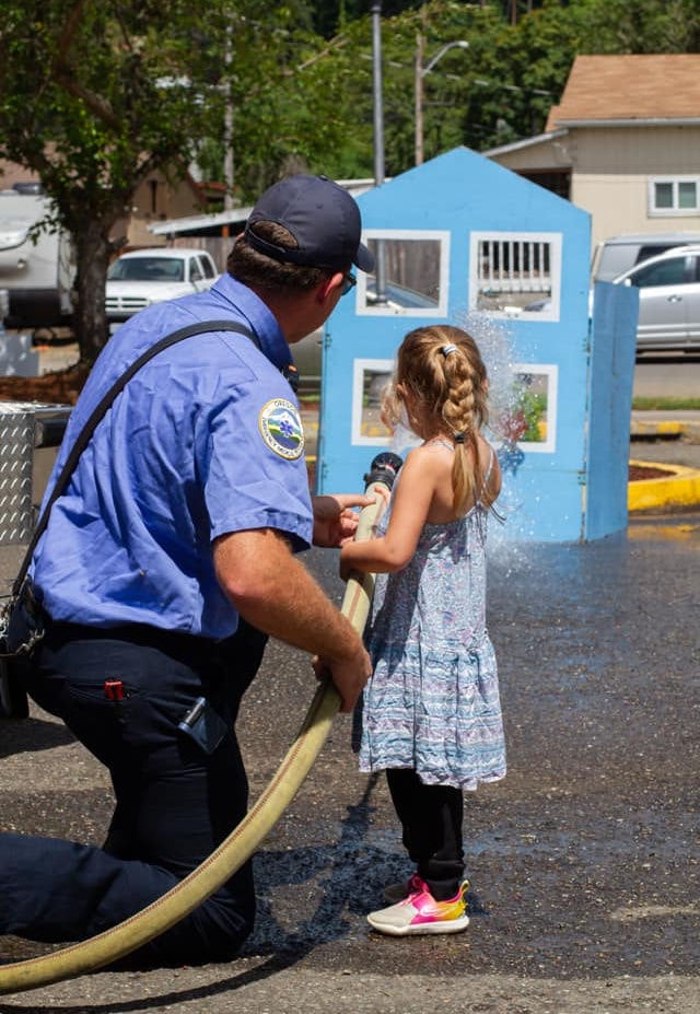 A firefighter kneels, helping a young girl hold a fire hose spraying water, in daylight with a blue playhouse in the background.