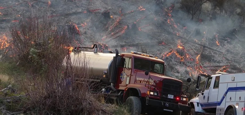 Firefighting trucks near a smoldering field with visible flames.