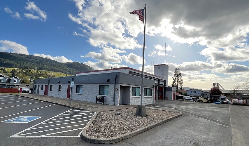 A single-story building, flagpole with US flag, parking lot with handicap spaces, in a hilly area, part cloudy sky.