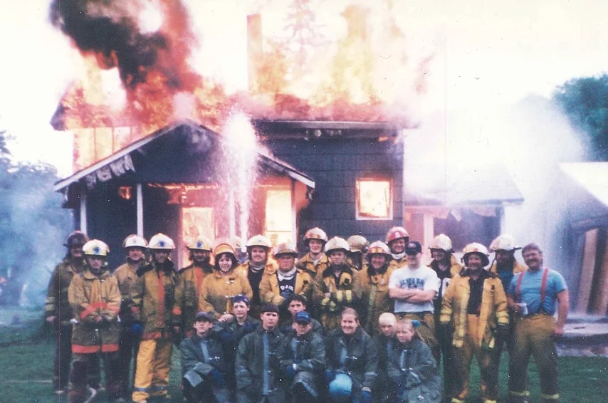 Firefighters pose in front of a burning house, likely during a training exercise.