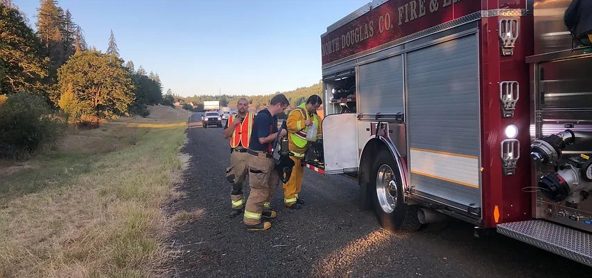 Firefighters beside a fire engine on a roadside, with traffic in the background.