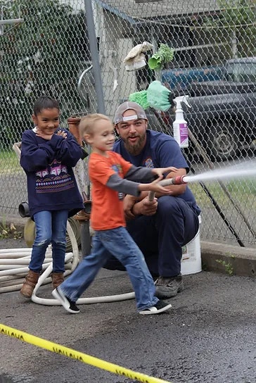 Firefighter helping a kid use a fire hose, another child watching. They're smiling and having fun.
